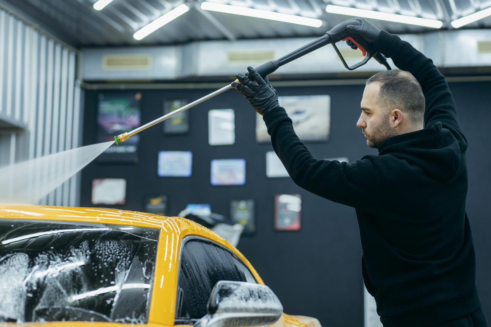 Um profissional enxágua um carro amarelo com uma lança de pressão em um box de lavagem.