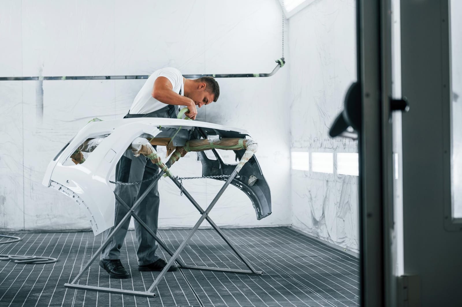 Um pintor protegendo um para-choque de carro sobre um suporte dentro de uma cabine de pintura.